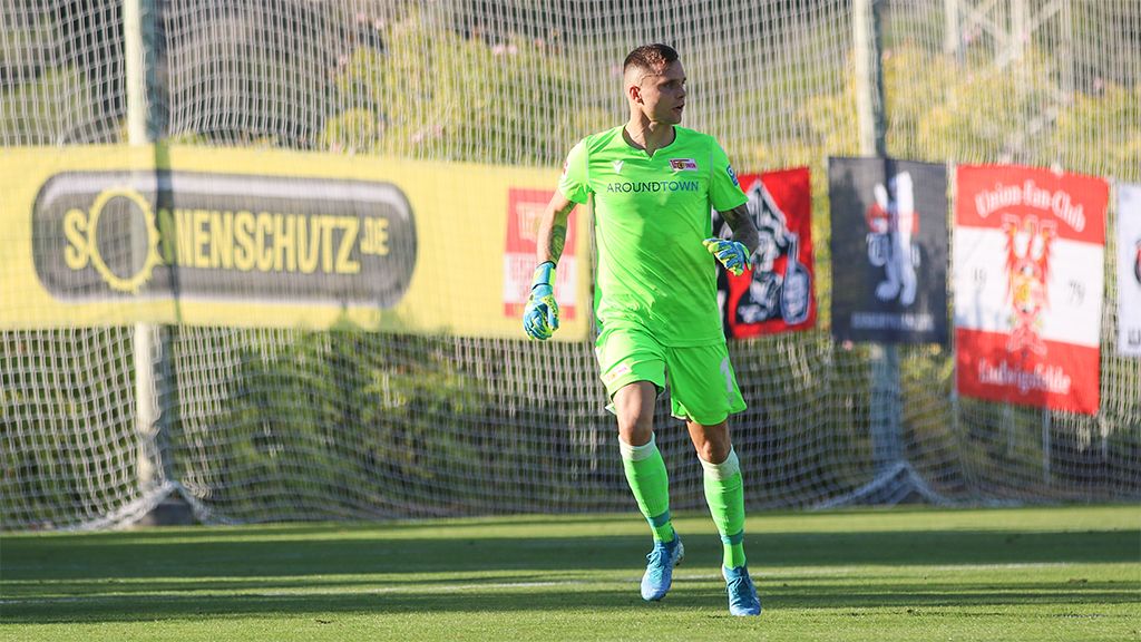 Ein Torwart in neonfarbener Ausrüstung steht auf dem Spielfeld, umgeben von Fußballnetz und Vereinsflaggen.