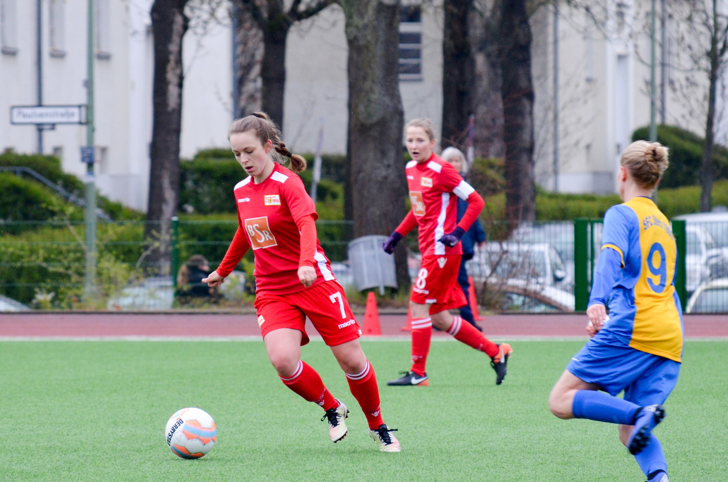 Eine Fußballspielerin in rotem Trikot dribbelt den Ball auf einem Grünfeld, während zwei Mitspielerinnen in gelben und blauen Trikots beobachten.