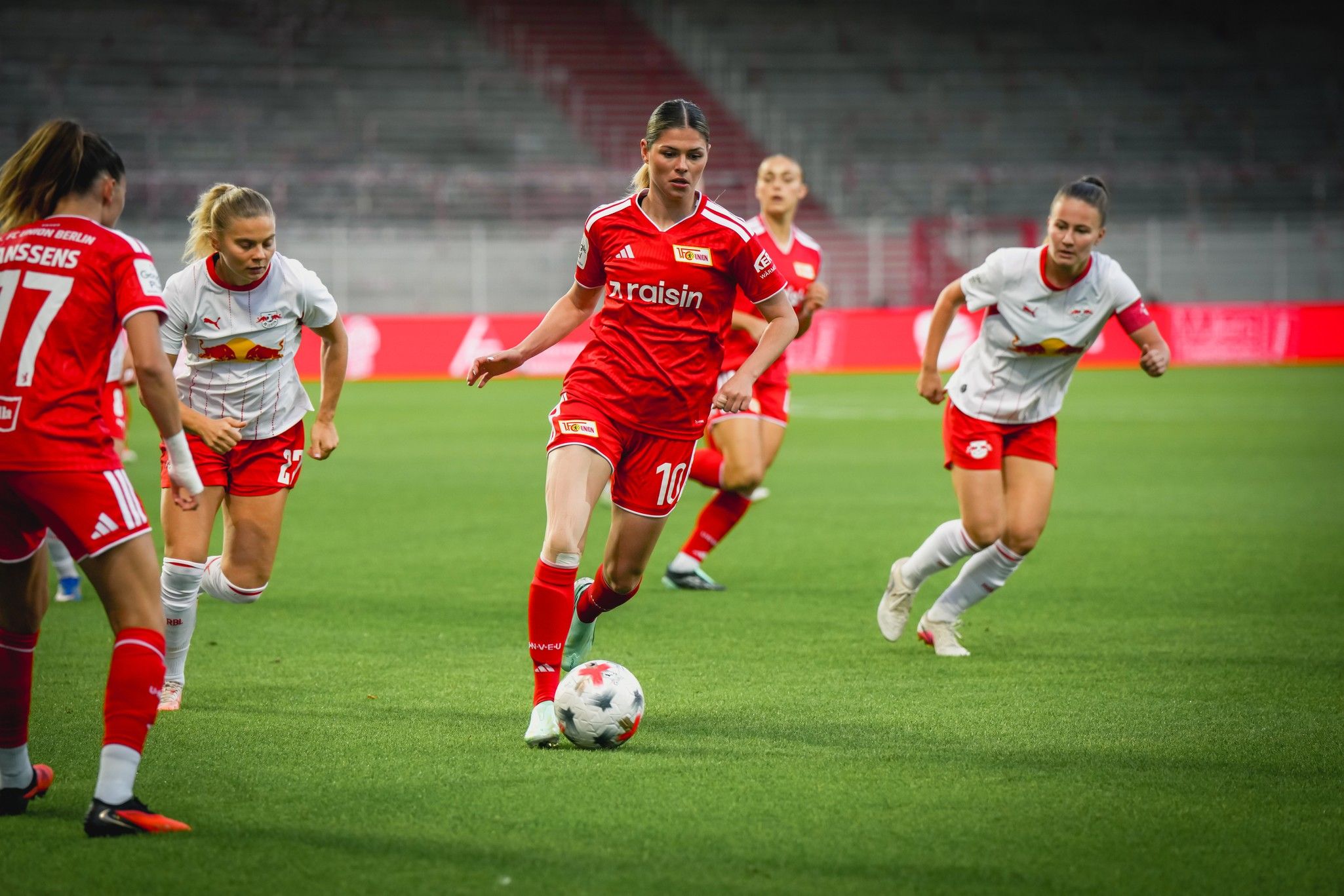 Eine Fußballspielerin in rotem Trikot dribbelt den Ball, während andere Spielerinnen in weißen Trikots sie verfolgen.