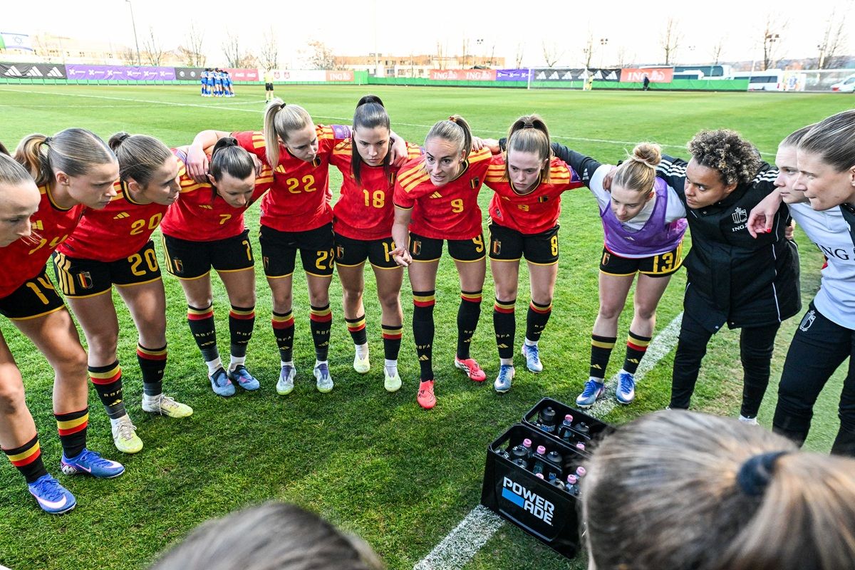 Eine Gruppe von Frauen in roten Fußballtrikots versammelt sich im Kreis auf einem Rasenplatz, während eine Trainerin spricht.