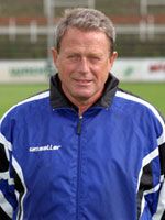 Ein Mann in einem blauen Sportanzug steht lächelnd auf einem Fußballfeld.