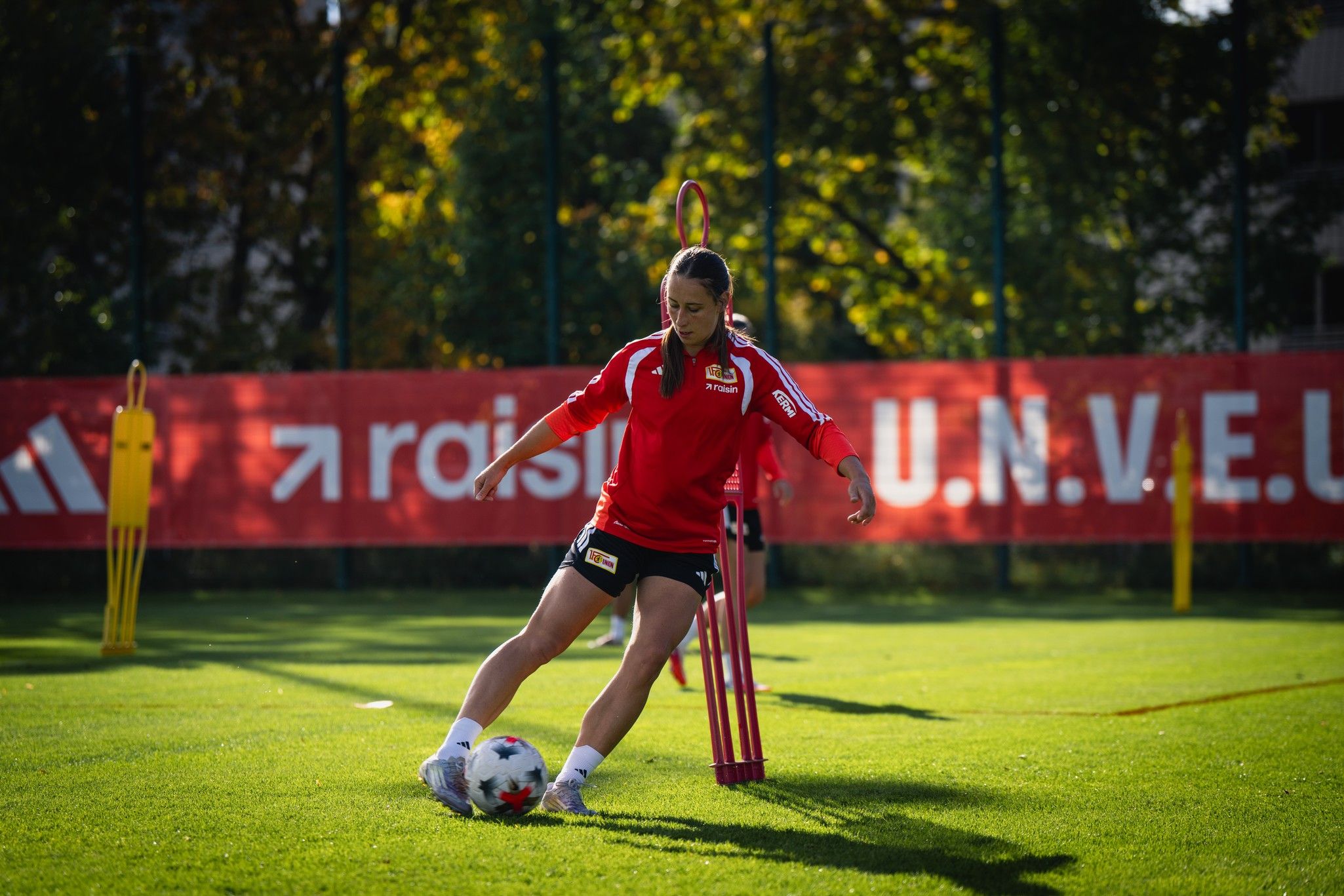 Eine Fußballspielerin in rotem Trikot trainiert auf einem Platz mit Übungsmarkierungen und Toren im Hintergrund.