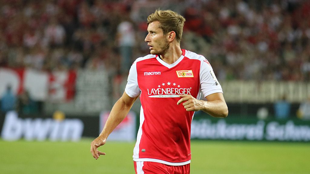 Soccer player in red jersey with white details on the field, spectators in the background.