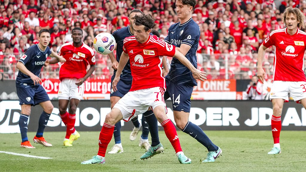 Spielszene mit zwei Fußballspielern im Wettkampf um den Ball, im Hintergrund jubelnde Fans in roten Trikots.