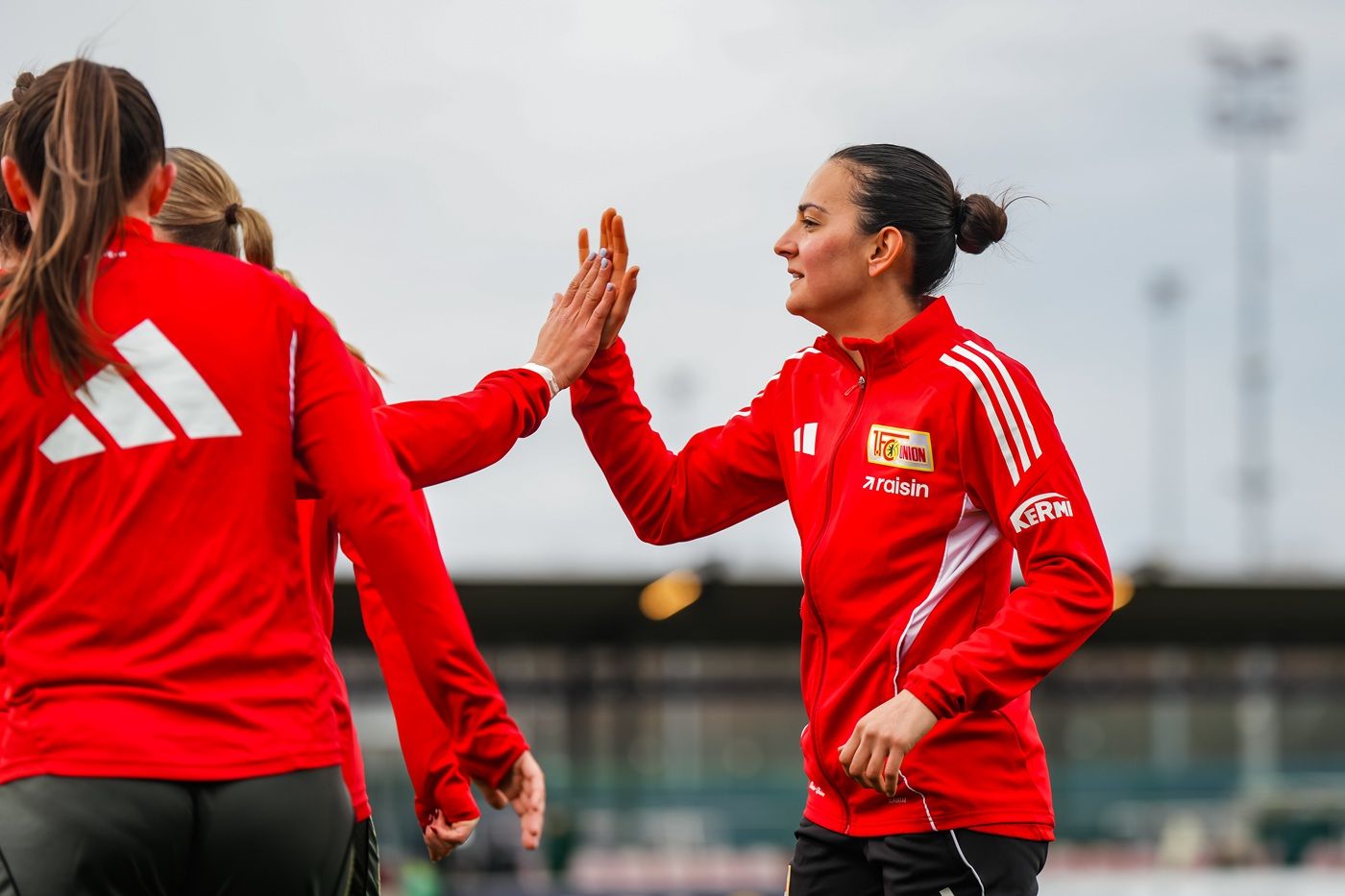 Frauenfußballmannschaft in roten Trikots, die sich beim Aufwärmen gegenseitig abklatscht. Wolkiger Himmel im Hintergrund.