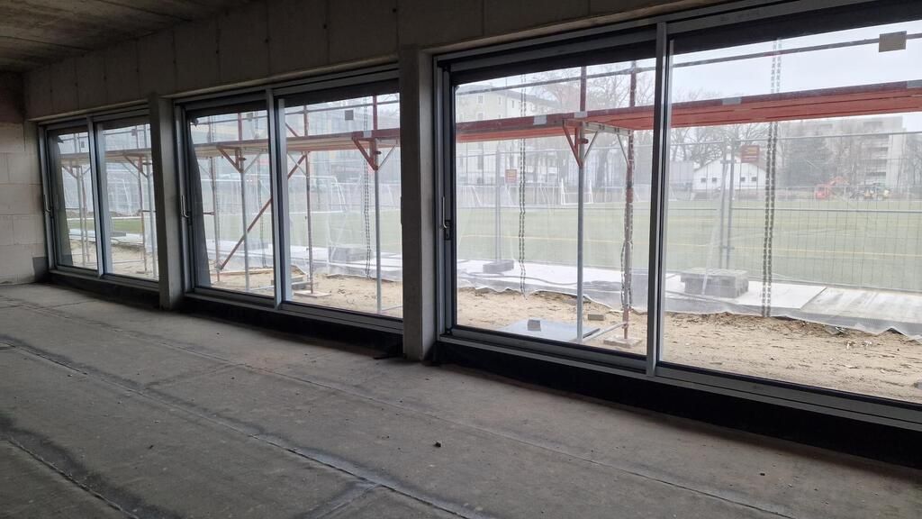 Blick durch große Fenster auf ein unfertiges Fußballfeld mit Baustellenmaterial im Vordergrund.