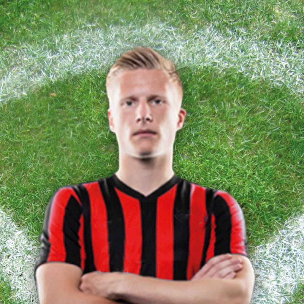 Ein junger Fußballspieler mit blondem Haar in einem rot-schwarzen Trikot steht mit verschränkten Armen auf einem Fußballfeld.