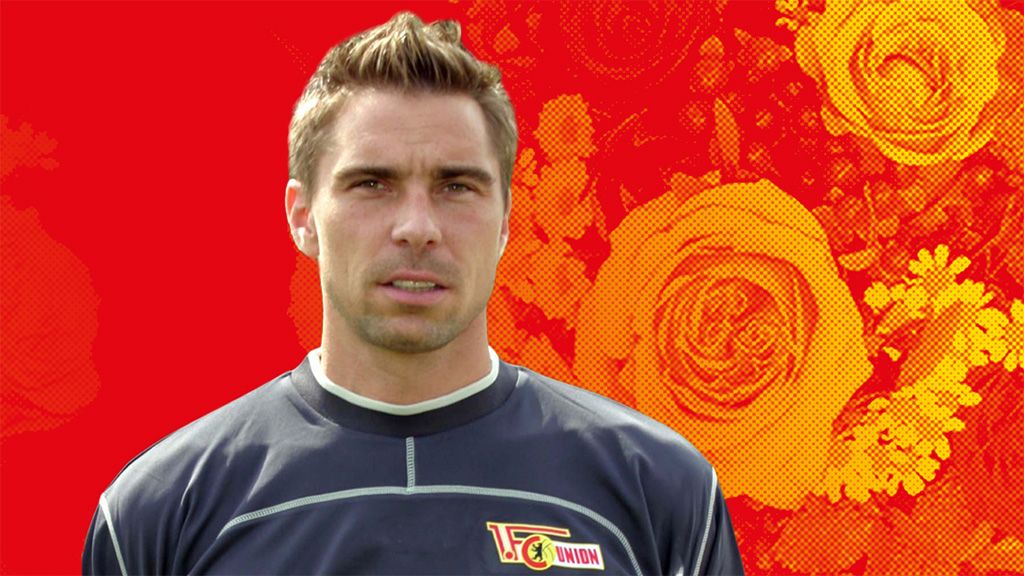 Ein Fußballspieler in einem grauen Trikot steht vor einem roten Hintergrund mit Blumenmustern.