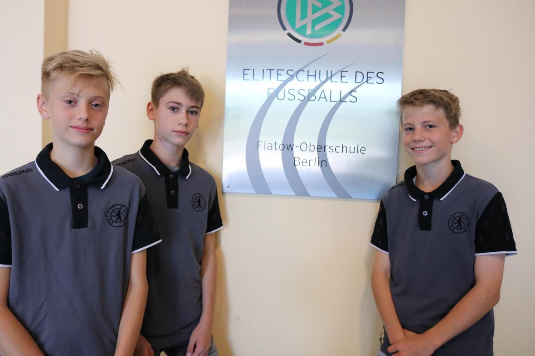 Drei Jugendliche in grauen Poloshirts stehen neben einem Schild mit der Aufschrift "Eliteschule des Fußballs, Flatow-Oberschule Berlin".