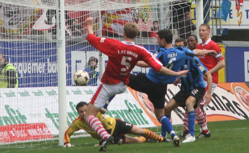 Ein Fußballspieler schießt den Ball ins Tor, während ein Verteidiger versucht, ihn abzuhalten. Die Torlinie und Zuschauer sind sichtbar.