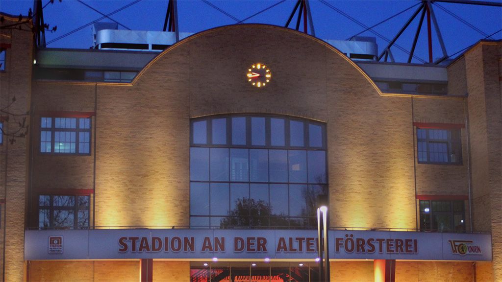 The picture shows the front view of the stadium at Alte Försterei, illuminated at dusk with a clock on the facade.