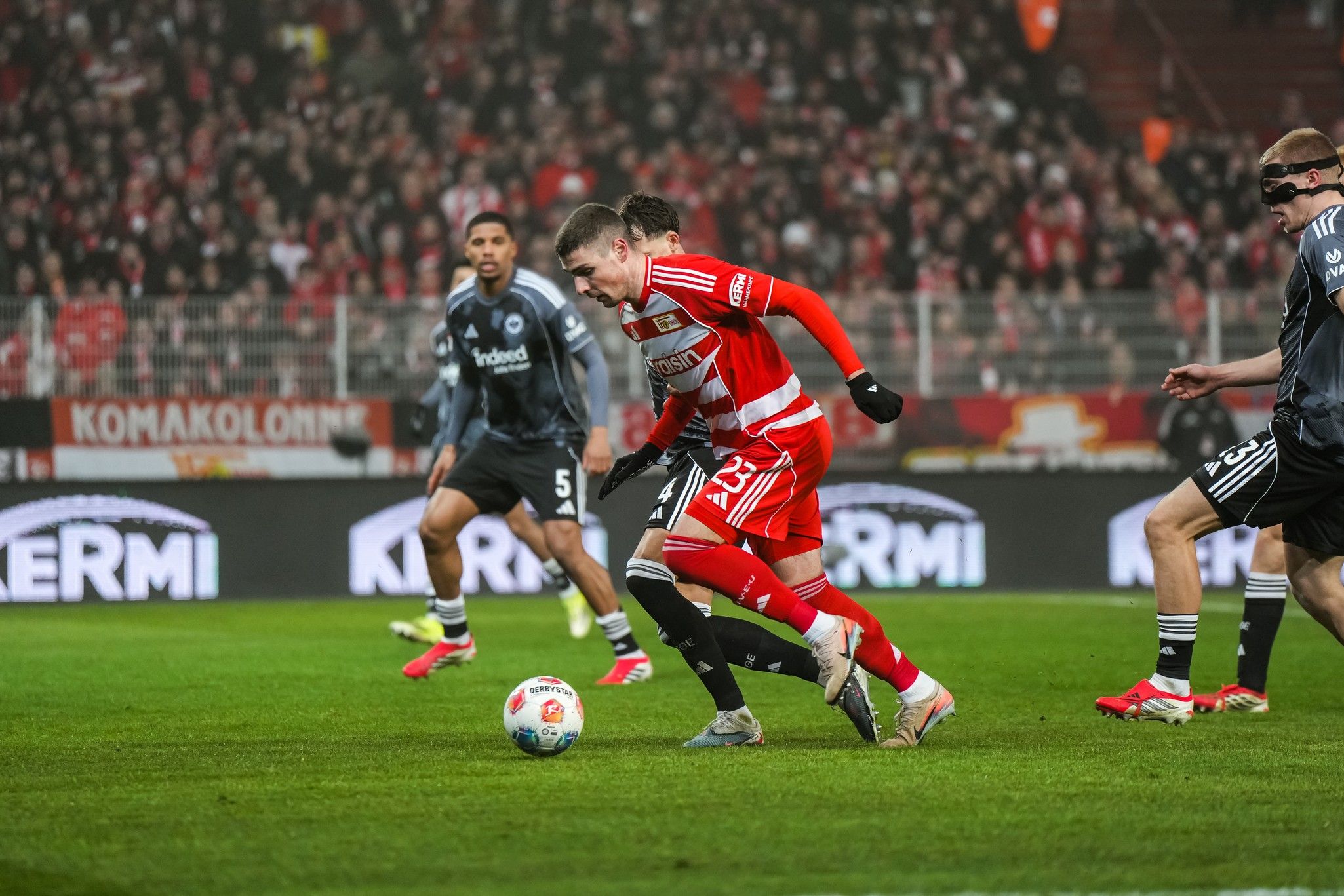 Ein Fußballspieler im roten Trikot dribbelt den Ball, während Gegner in dunklen Trikots nahe sind. Zuschauer im Hintergrund.