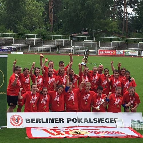 Gruppensiegerehrung eines Jugendfußballteams in roten Trikots mit Pokal und Medaillen auf einem Sportgelände.