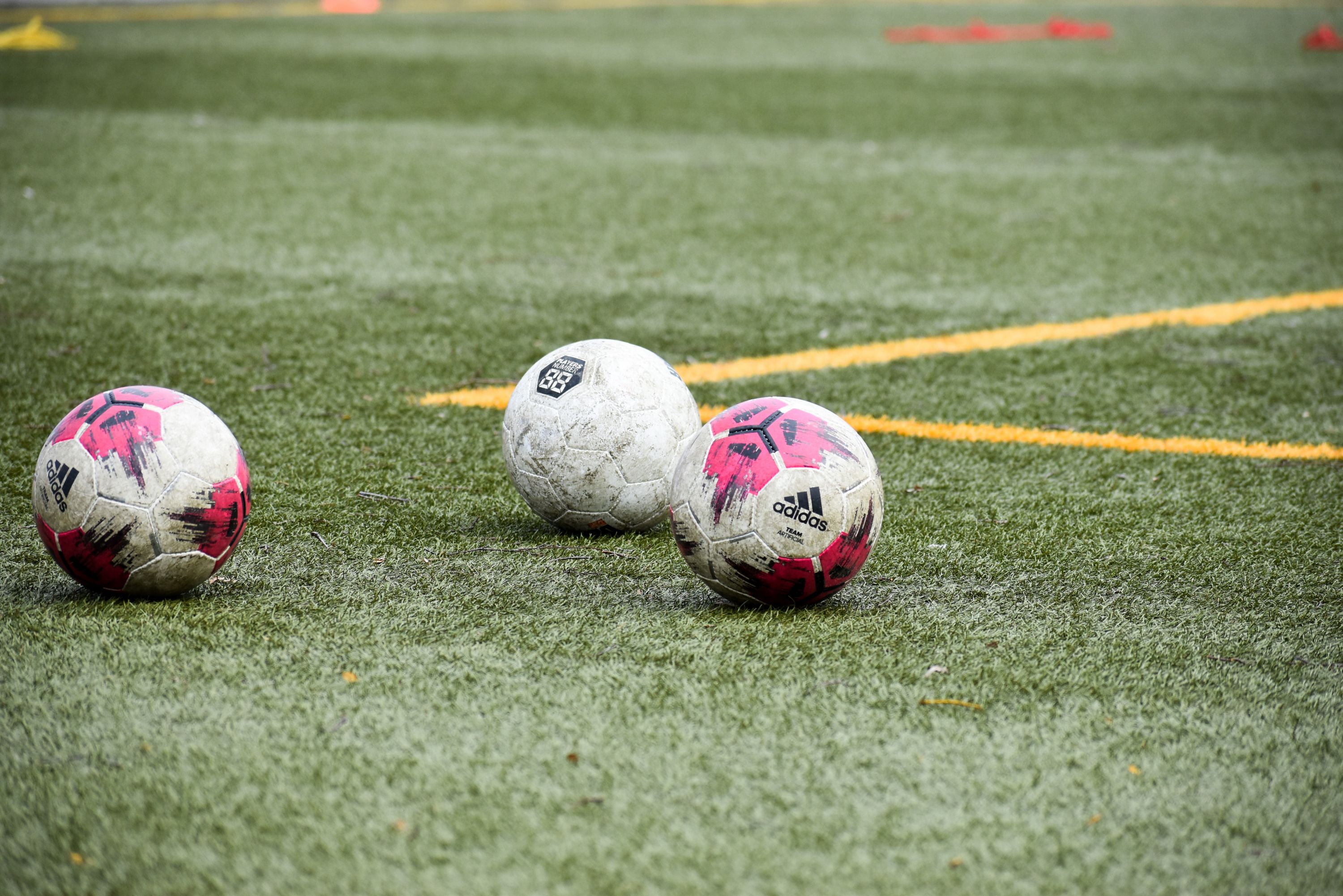 Drei Fußball mit verschiedenen Designs liegen auf einem Rasenfeld mit Markierungen.