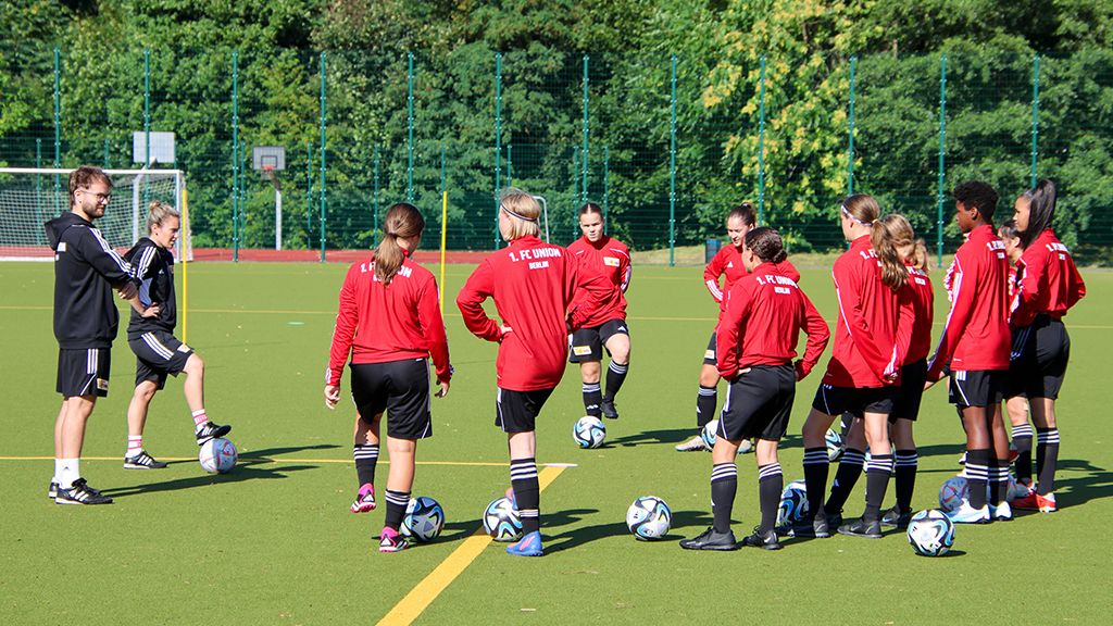 Mädchenfußballmannschaft in roten Trikots trainiert auf einem Platz mit mehreren Fußbällen und einem Trainer.