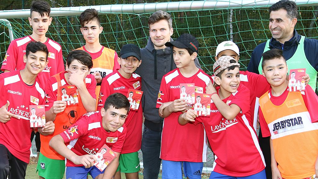 Gruppe von fußballspielenden Jungen in roten Trikots posiert lächelnd vor einem Tor, mit einer Person im Hintergrund.