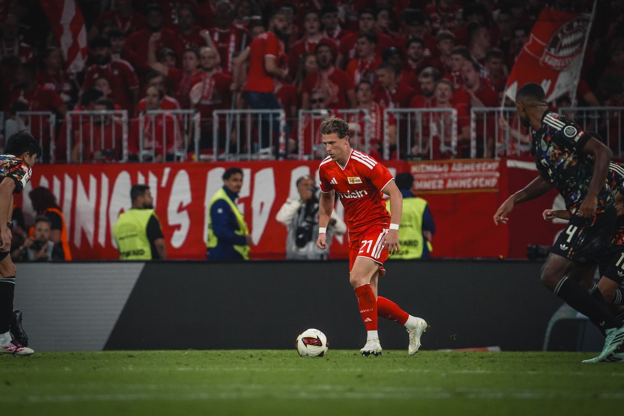 Ein Fußballspieler in rotem Trikot dribbelt den Ball, während Fans im Hintergrund jubeln und Banner zeigen.