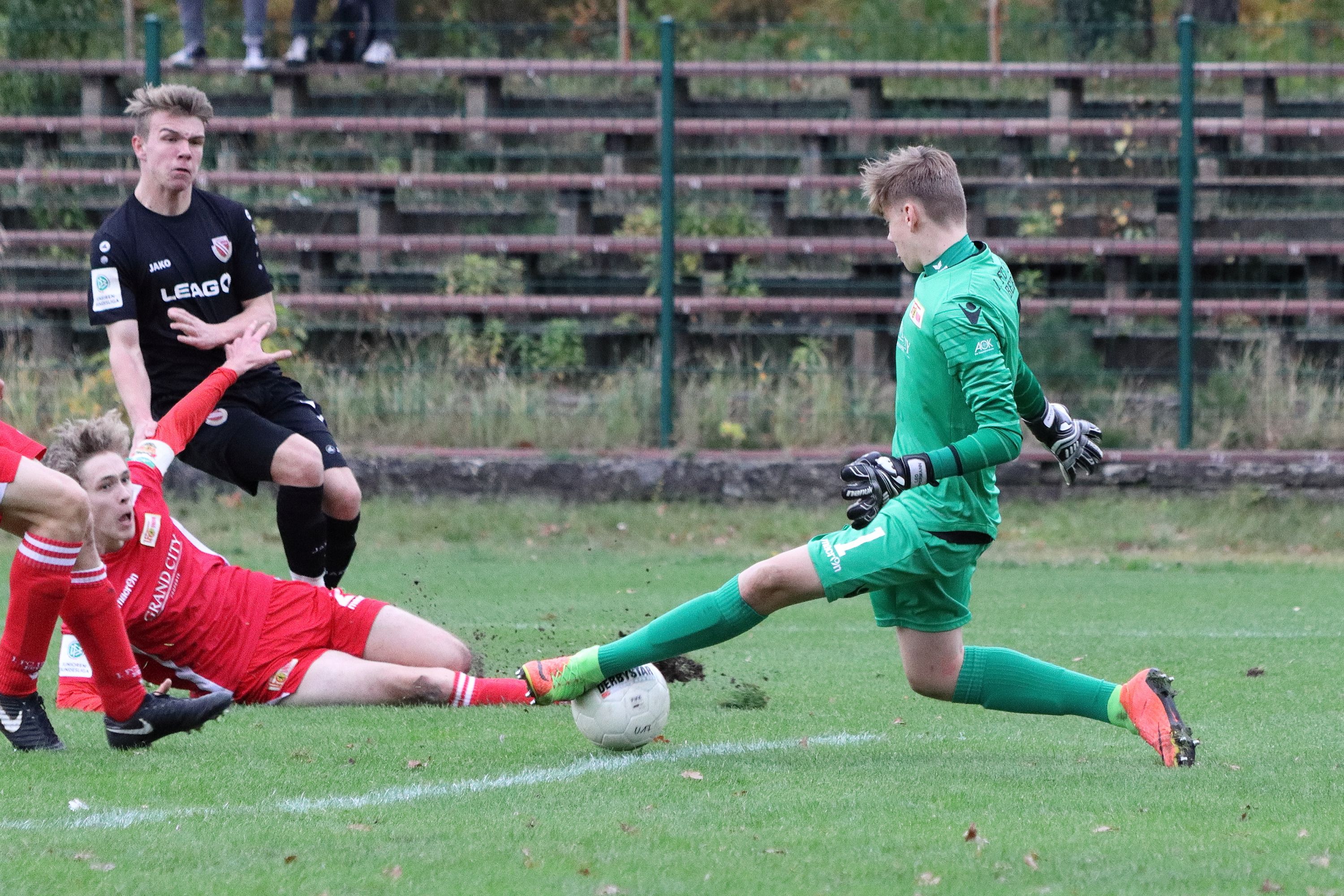 Ein Torwart in grüner Ausrüstung pariert einen Ball, während ein Spieler in roter Ausrüstung am Boden liegt. Im Hintergrund sind Zuschauerplätze zu sehen.