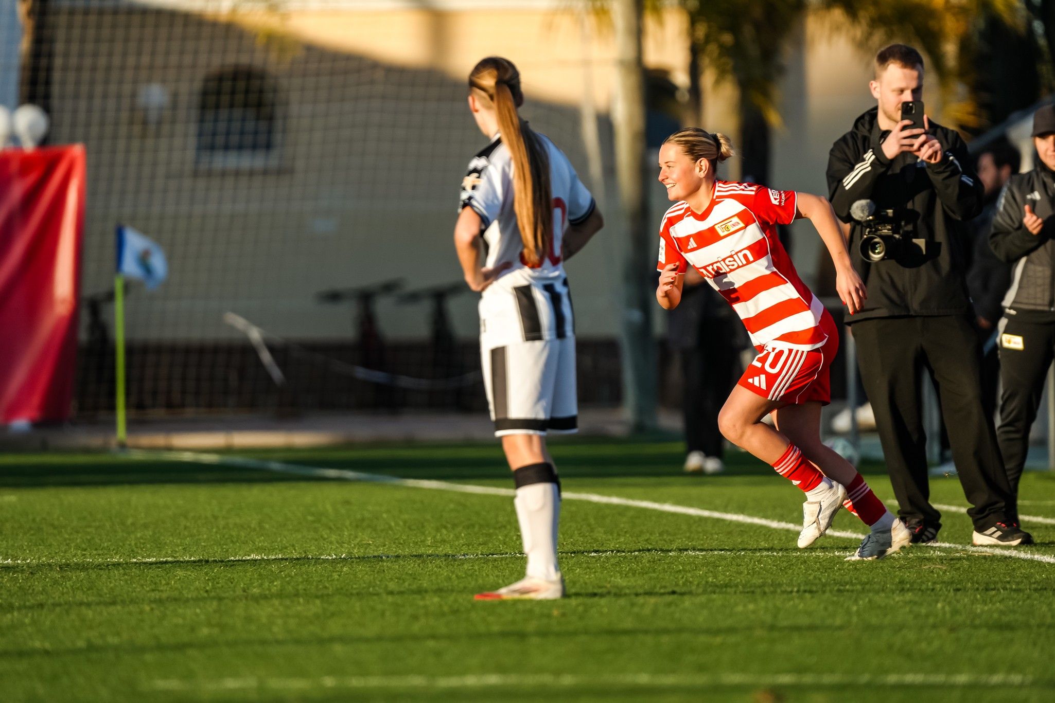 Eine Spielerin in rot-weißem Trikot feiert einen Torerfolg, während eine gegnerische Spielerin in schwarz-weißer Uniform zuschaut.