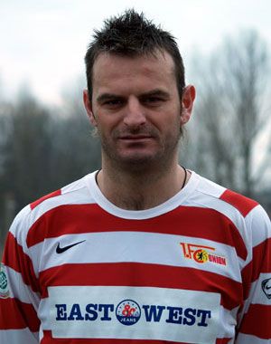 Ein Fußballspieler trägt ein rot-weiß gestreiftes Trikot mit dem Logo eines Vereins und steht im Freien vor unscharfen Hintergrund.