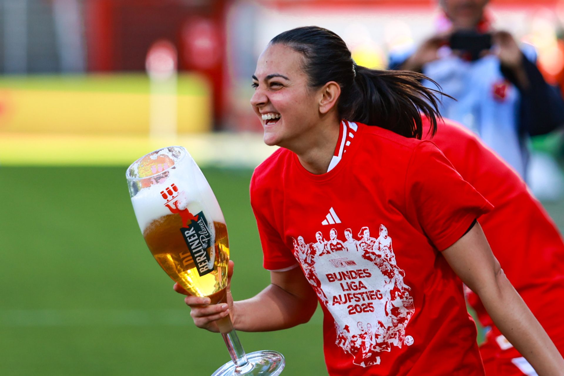Eine fröhliche Spielerin in einem roten T-Shirt hält einen großen Bierkrug und feiert den Aufstieg in die Bundesliga.