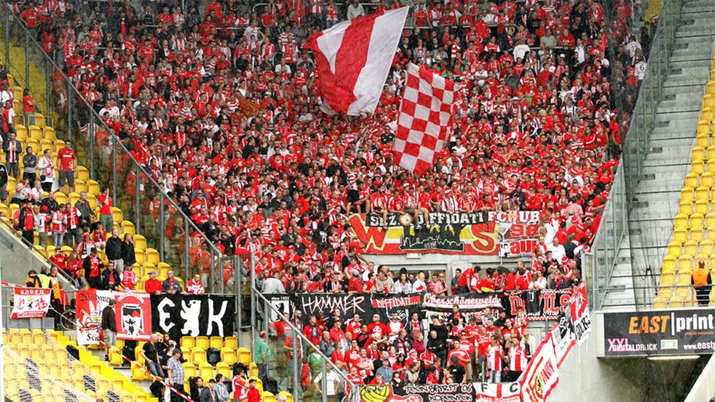 Vereinsfans in roter und weißer Kleidung feiern in einem Stadion, umgeben von Schildern und Fahnen.