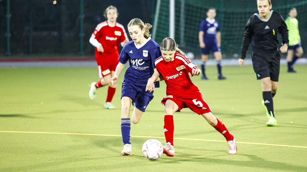 Zwei Mädchen spielen Fußball auf einem Kunstrasenfeld, eines in blauer und das andere in roter Teamkleidung.