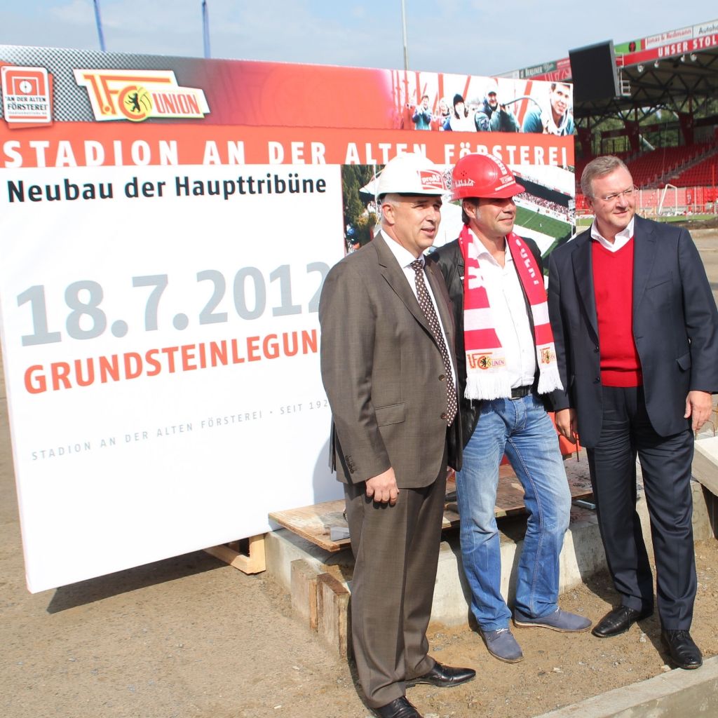 Drei Männer stehen vor einem Schild, das den Neubau der Haupttribüne des Stadions an der Alten Försterei ankündigt.