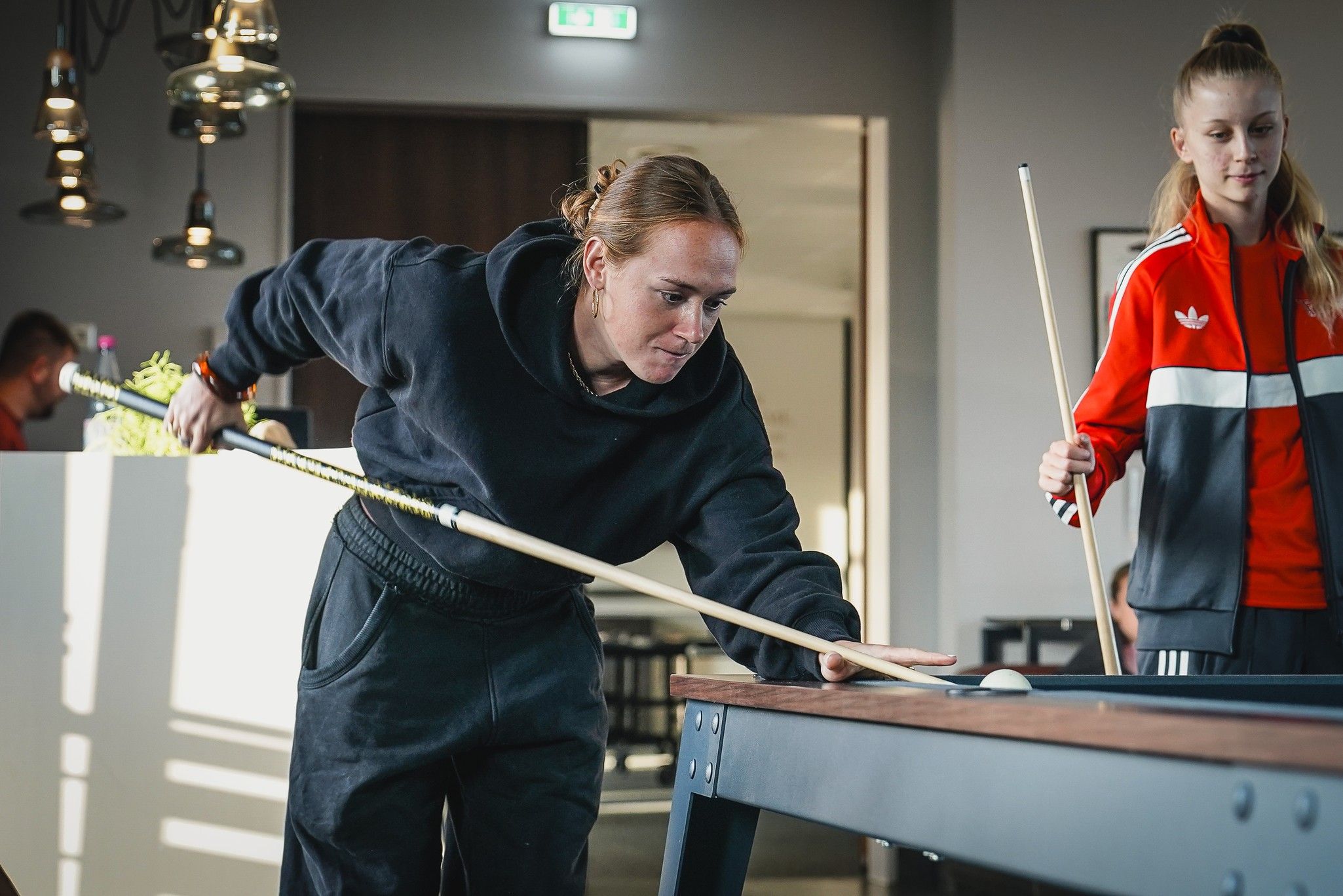 Eine Frau spielt Billard, während eine zweite Person beobachtet. Beide tragen sportliche Kleidung in einem modernen Raum.