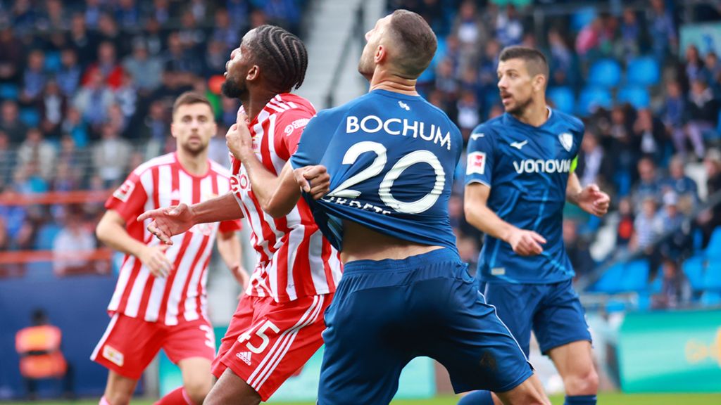 Zwei Fußballspieler im Wettkampf um den Ball während eines Spiels in einem Stadion. Zuschauer im Hintergrund.