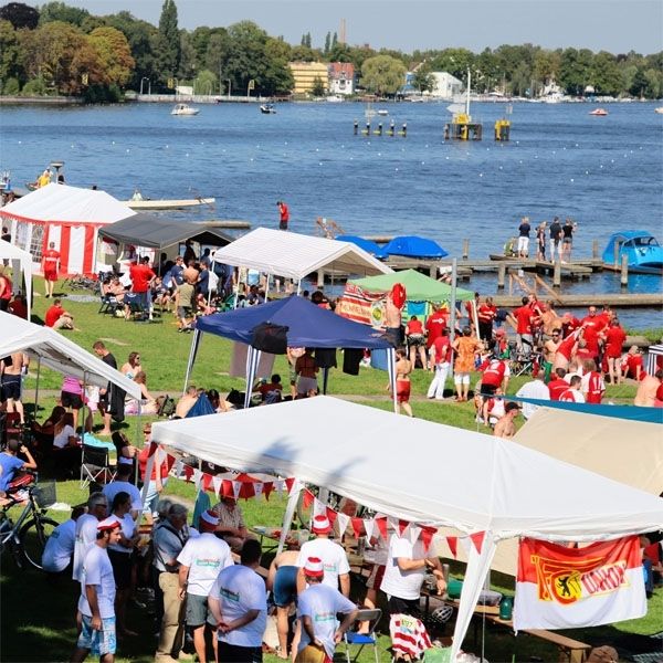 Menschen in Festkleidung feiern am Wasser, umgeben von Zelten und Bänken, mit Blick auf eine ruhige Wasseroberfläche.
