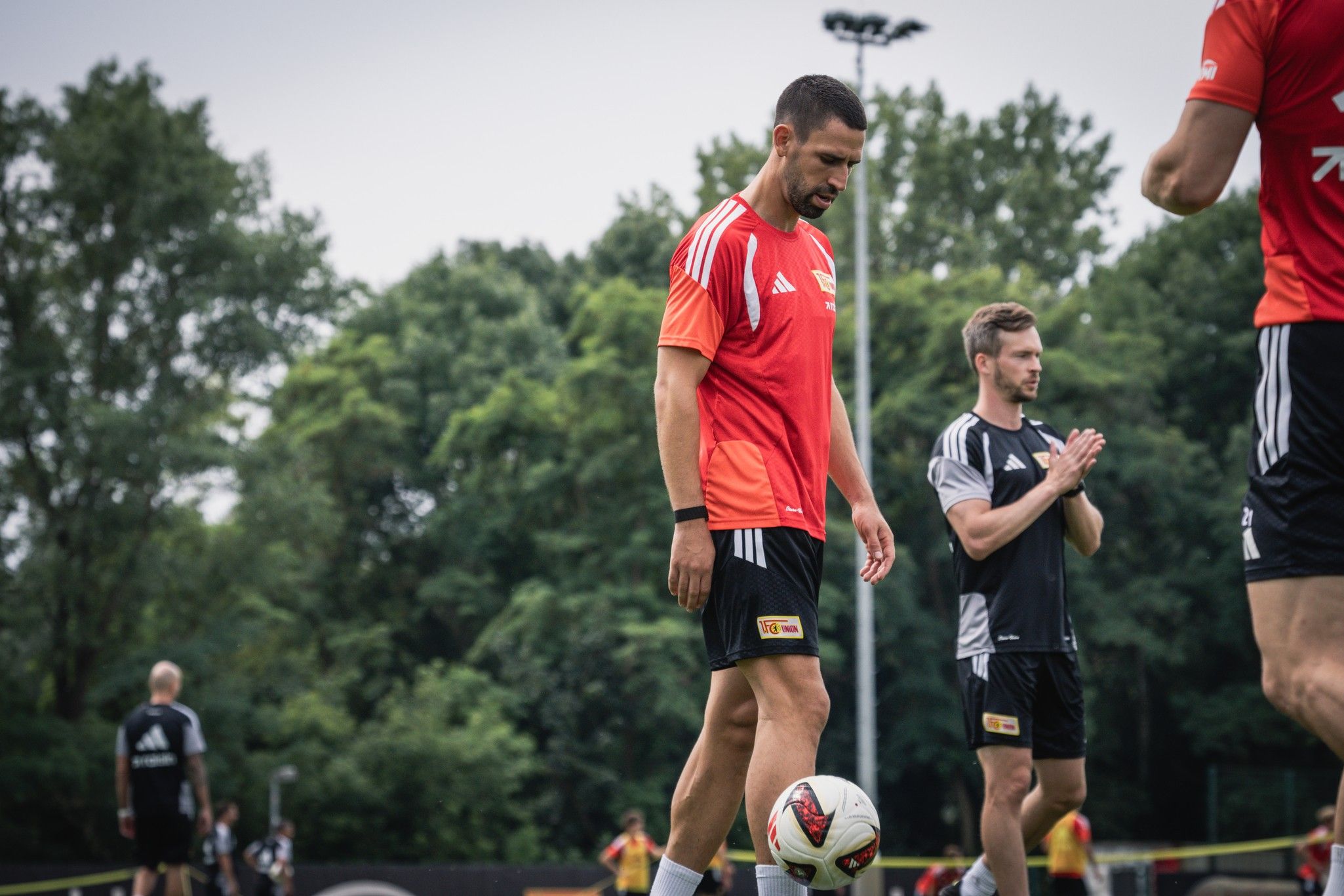 Fußballspieler trainieren auf dem Platz, einer jongliert einen Ball, während andere Übungen durchführen. Grüne Bäume im Hintergrund.