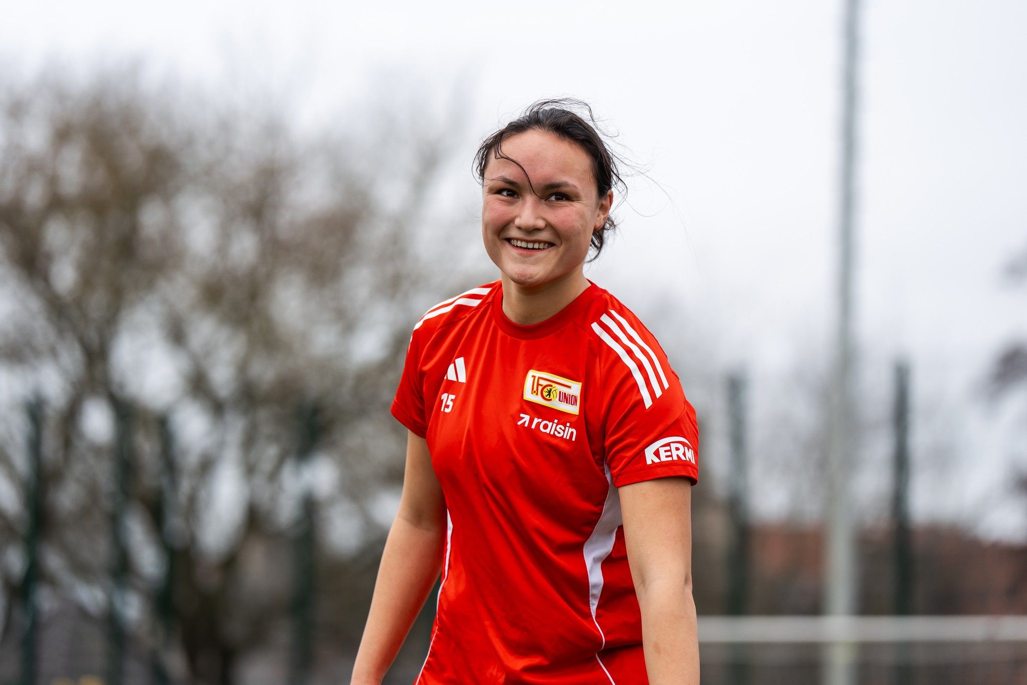 Eine lächelnde Spielerin in einem roten Trikot steht auf einem Fußballplatz, im Hintergrund unscharfe Bäume und ein Fußballtor.