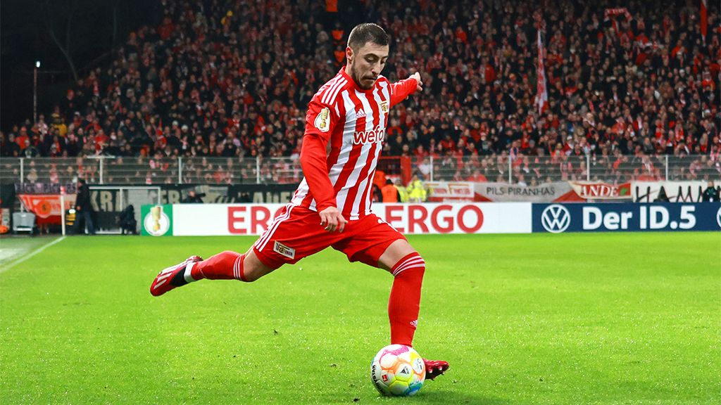 A player in red and white jerseys kicks a soccer ball on a stadium field, surrounded by cheering fans.