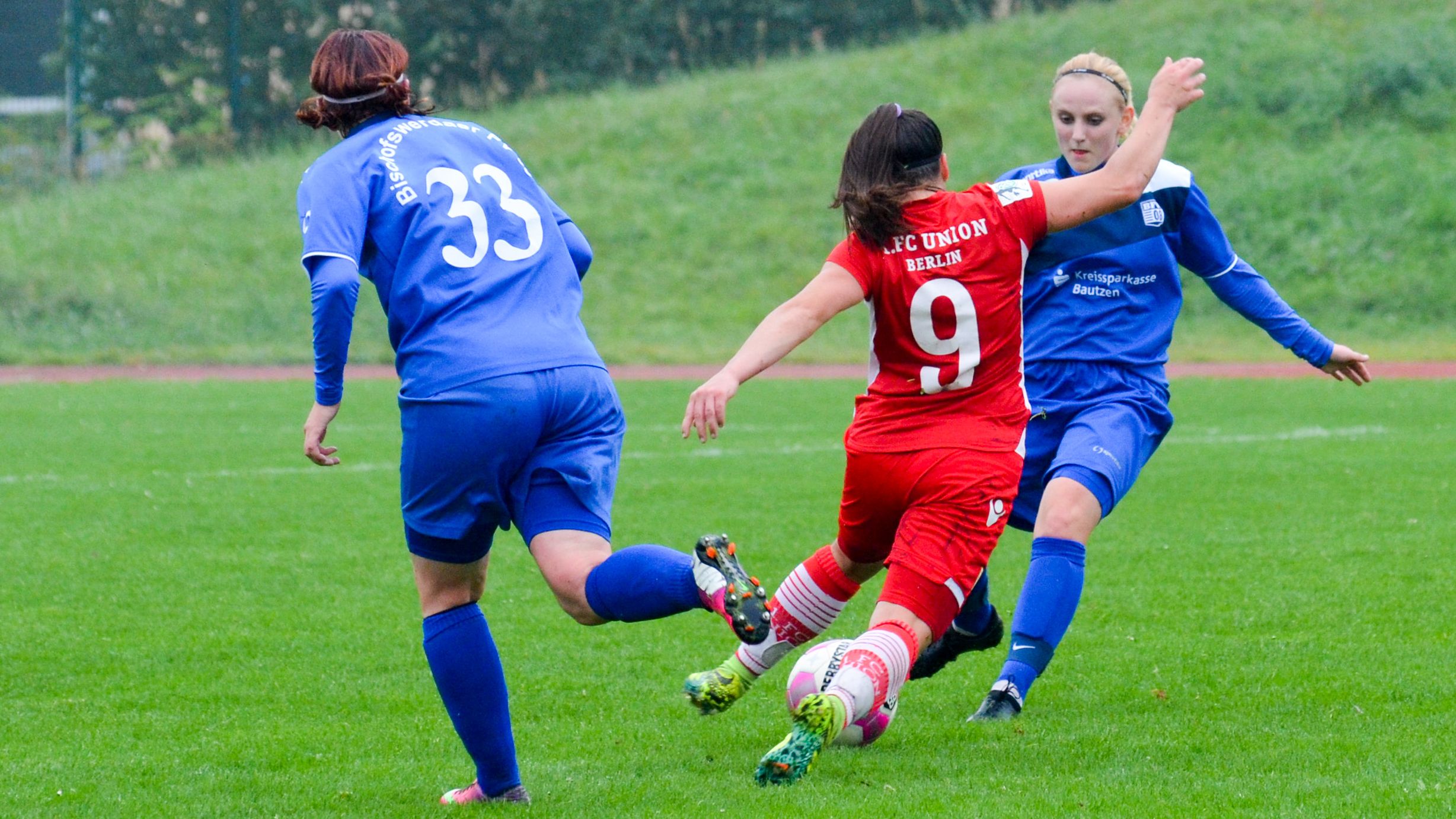 Zwei Spielerinnen in blauen und roten Trikots kämpfen um den Ball auf einem grünen Fußballfeld.