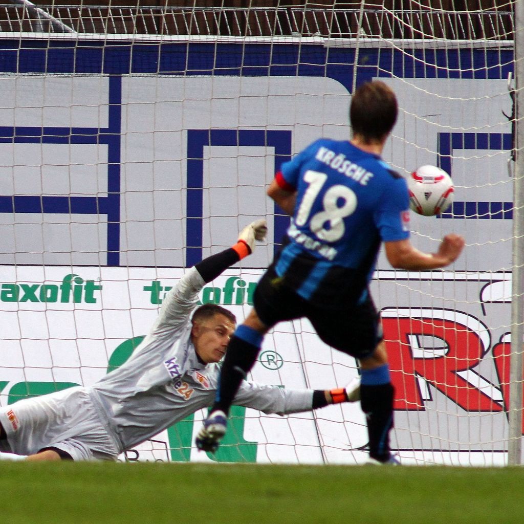 Ein Torwart springt, um einen Schuss auf das Tor zu parieren, während ein Spieler bereit ist, den Ball zu schießen.