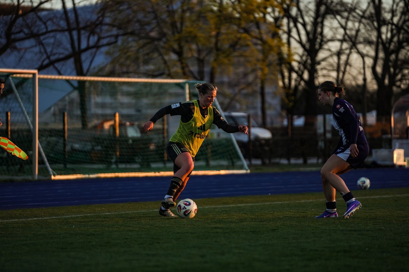 Eine Spielerin in gelber Trainingskleidung dribbelt den Ball auf einem Fußballfeld bei Sonnenuntergang.