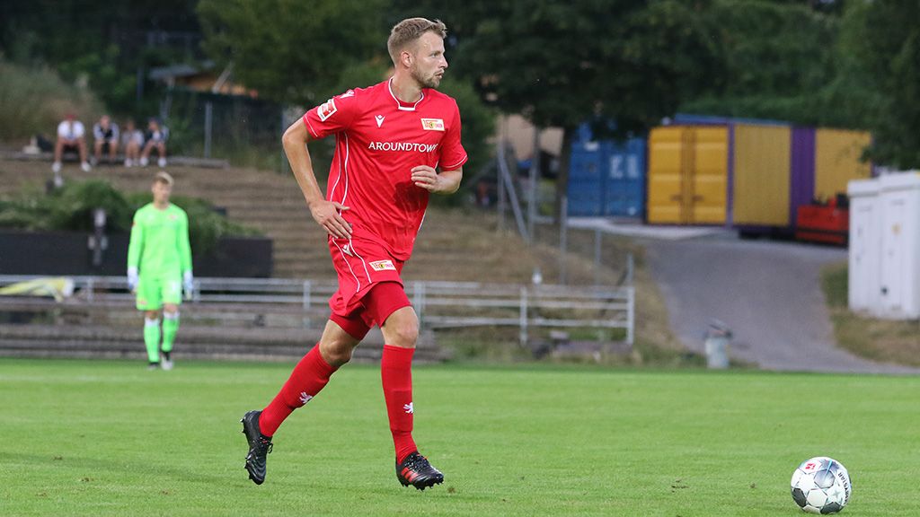 A soccer player in red gear runs across the field, while a goalkeeper stands in the distance.