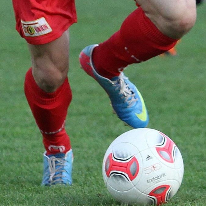 Ein Spieler in roter Sportkleidung steht auf einem grünen Fußballfeld und tritt einen weißen Fußball.