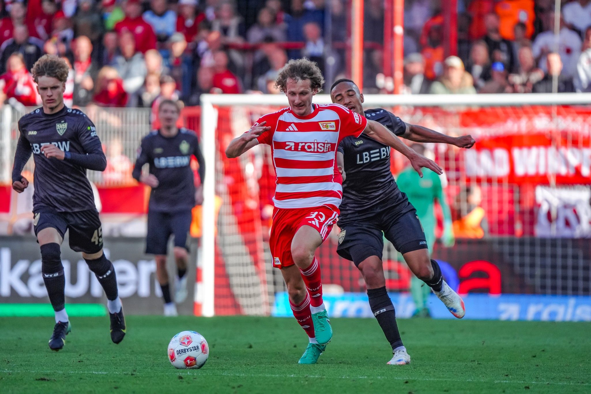 Spielszene mit einem Spieler in rotem Trikot, der von einem Verteidiger in dunklem Trikot verfolgt wird. Fans im Hintergrund.