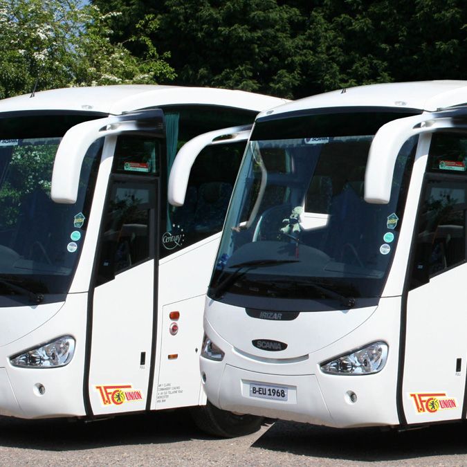 Zwei weiße Reisebusse stehen nebeneinander auf einem Parkplatz, umgeben von grünen Bäumen.