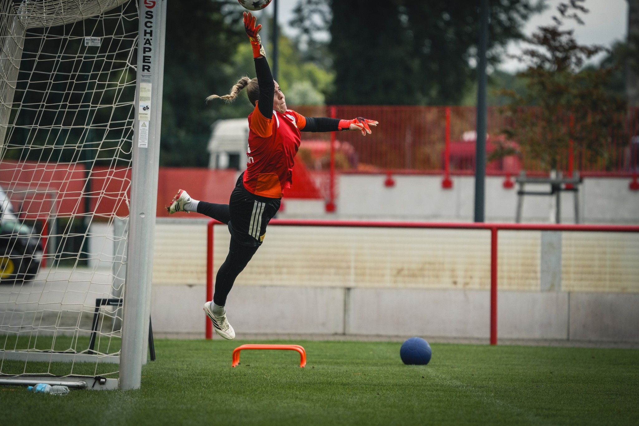 Torwart springt hoch, um einen Ball während des Trainings zu fangen, im Hintergrund ein Fußballfeld und ein Tor.