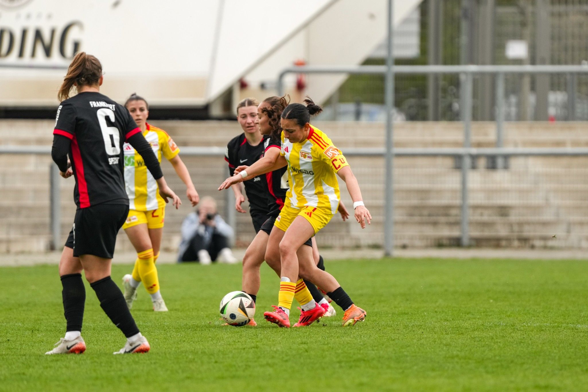 Zwei Fußballspielerinnen kämpfen um den Ball auf einem Rasenplatz, während andere Spielerinnen in der Nähe sind.