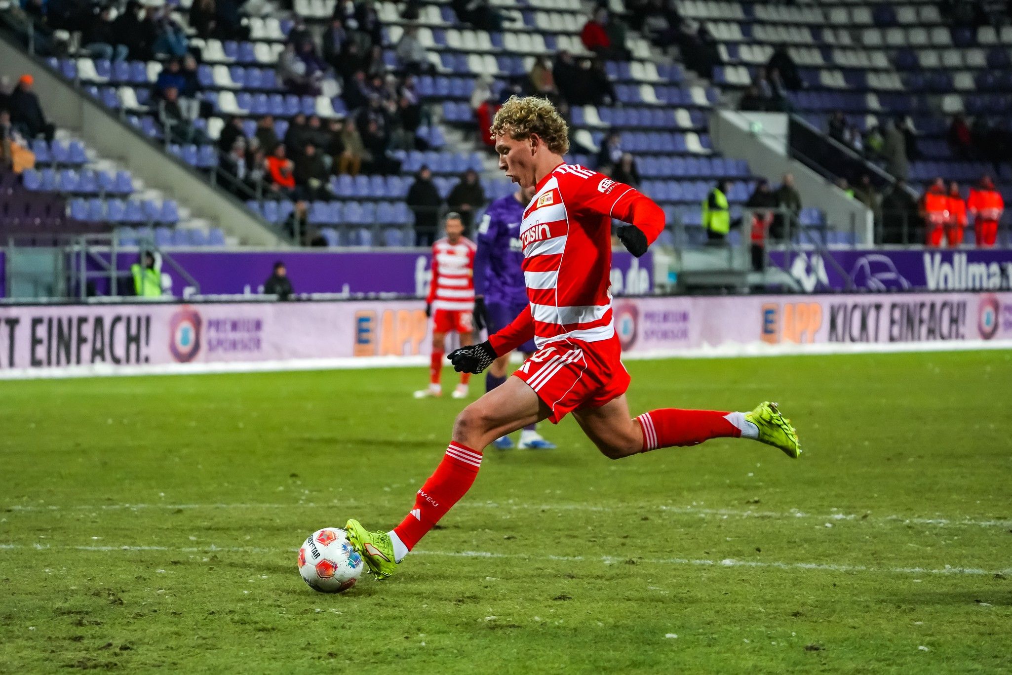 Spieler in rot-weißem Trikot schießt den Ball auf einem grünen Fußballplatz, umgeben von leeren Zuschauerplätzen.