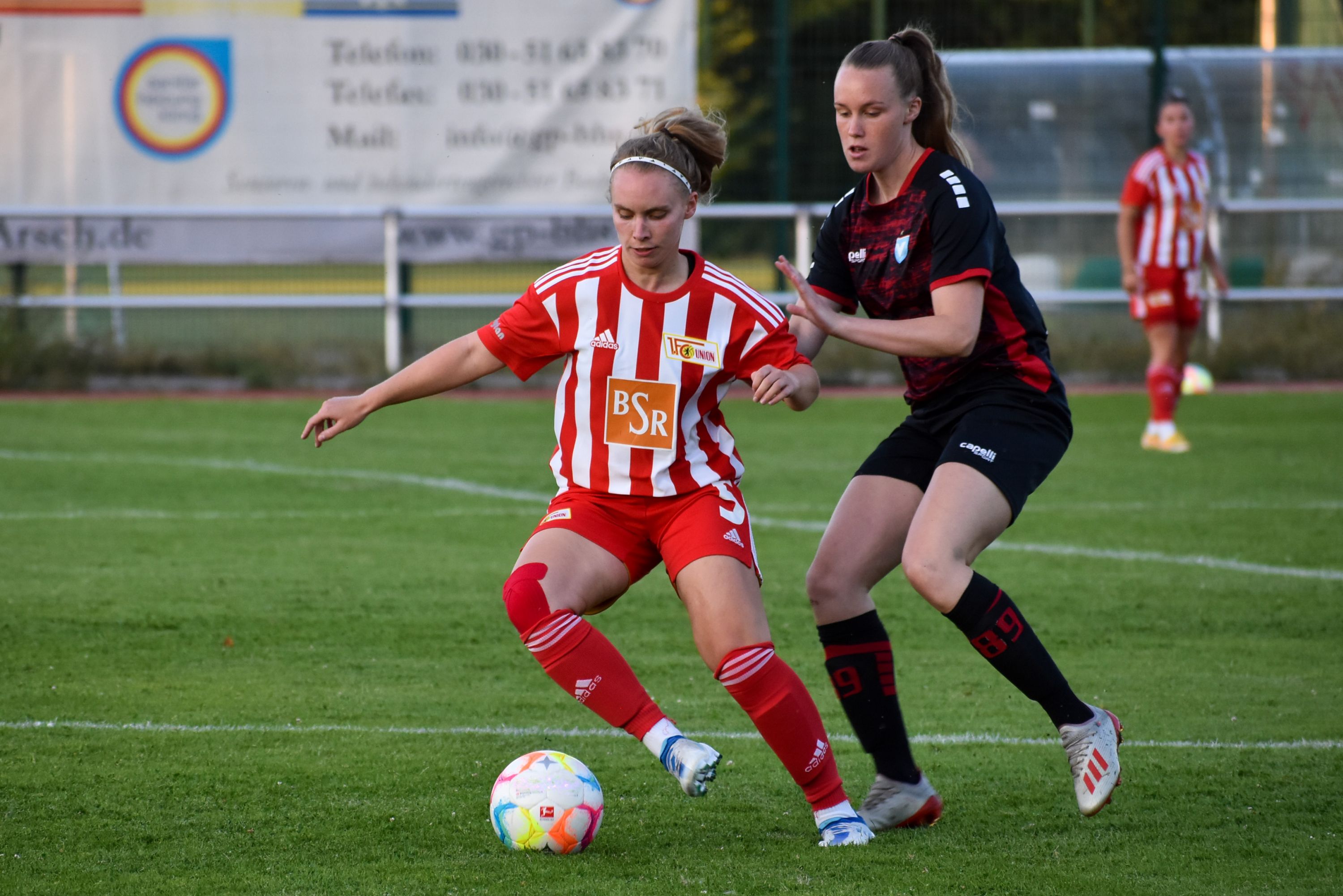 Spielerzwei Frauen im Fußballtrikot dribbelt den Ball, während die andere versucht, den Ball zu erobern. Rasenplatz im Hintergrund.