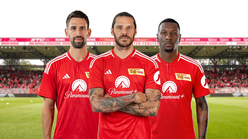 Three soccer players in red jerseys stand side by side in front of a stadium background, smiling and confident.