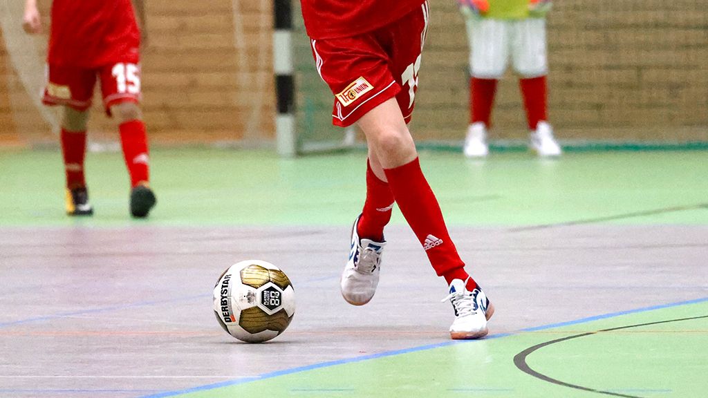 Ein Spieler in roter Sportkleidung dribbelt einen Fußball in einer Halle. Im Hintergrund sind weitere Spieler zu sehen.