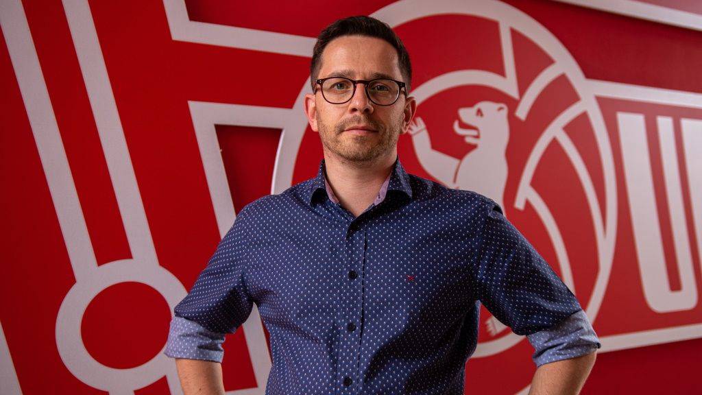 Mann mit Brille in blauem Hemd steht vor einer roten Wand mit einem Logo im Hintergrund.