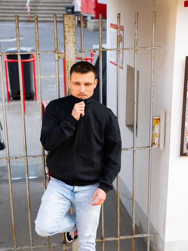 Junger Mann in schwarzem Sweatshirt steht an einem Gittertor und hält einen Klickverschluss in der Hand.