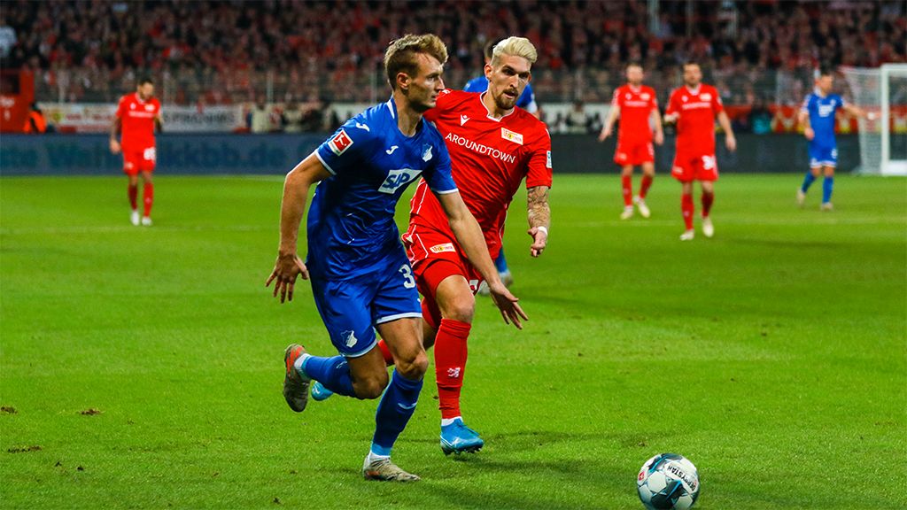 Two soccer players in a dynamic game situation on the field, the ball is between them. Spectators in the background.